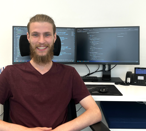 Lächelnder junger Mitarbeiter an einem Arbeitsplatz mit zwei Bildschirmen, die Code zeigen.